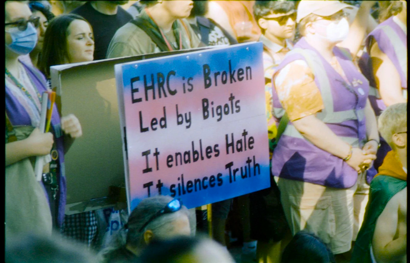 A photo from a Trans Rights Protest taken on film by Lily when live reporting for What The Trans. In a large group of people the centre of the picture is taken up by a sign saying "EHRC is Broken, Led by bigots, It enables hate, It silences Truth" the background of the sign is the trans flag colours of Pink Blue and White